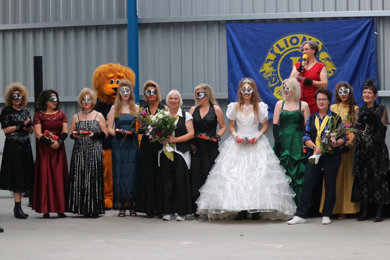 Zum Abschluss der Show Damen in Abendgarderobe mit Halbmasken vor der Bühne aufgereiht. Dazwischen die beiden Hauptorganisatorinnen mit je einem Blumenstrauß im Arm. Im Hintergrund überragt unser Löwen-Maskottchen Leo die Gruppe.