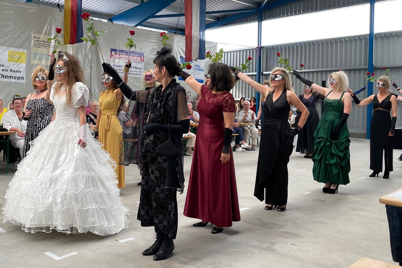 Damen in Abendkleidung stehen Spalier durch das eine junge Frau im Brautkleid schreitet.