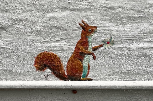 Ein aufgerichtetes Eichhörnchen hält ein Briefchen mit einem Herz in der Pfote. Es ist über einem dünnen Rohr auf eine Hauswand gemalt.