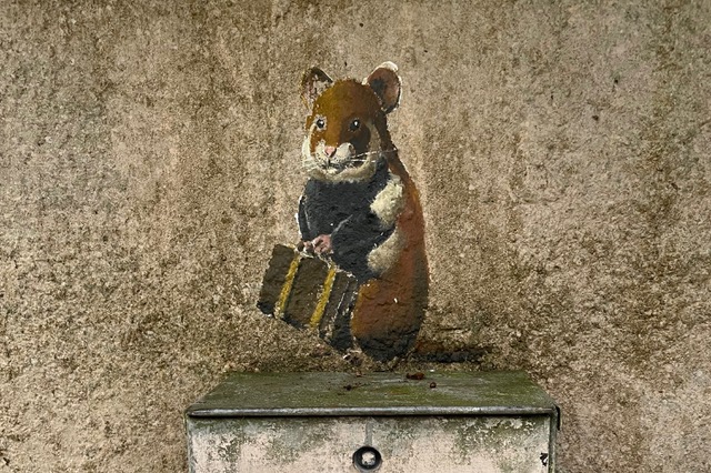 Ein stehender Feldhamster ist mit einem Koffer in der Pfote über einem Schacht auf eine Hauswand gemalt.