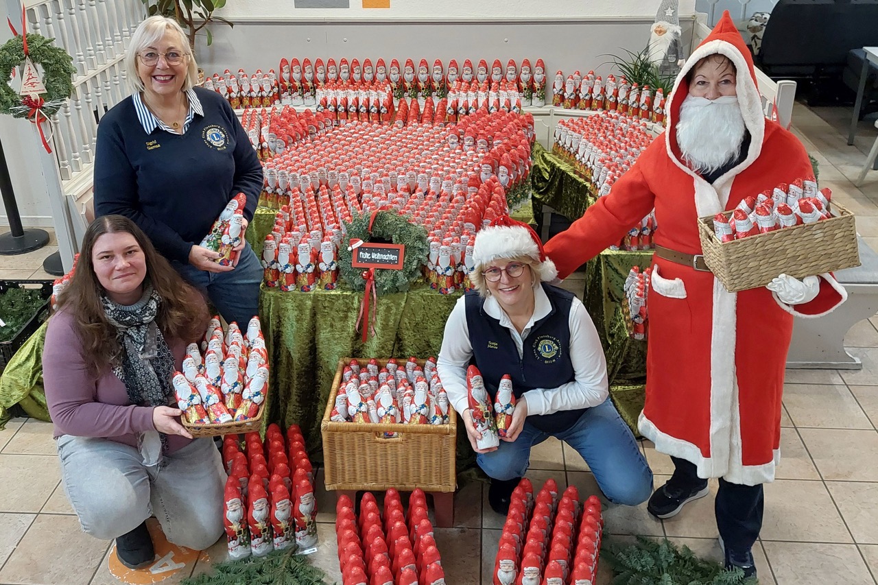 Die Präsidentin 2024-2025 sitzt mit zwei Schokoladen-Weihnachtsmännern in den Händen links von über 1000 Schoko-Männern, die auf Tischen und am Boden aufgestellt sind. Vor ihr die Leiterin der Tafel. Rechts auf dem Bild steht eine Person als Nikolaus verkleidet. Eine weitere Lionsfreundin hockt neben ihr.