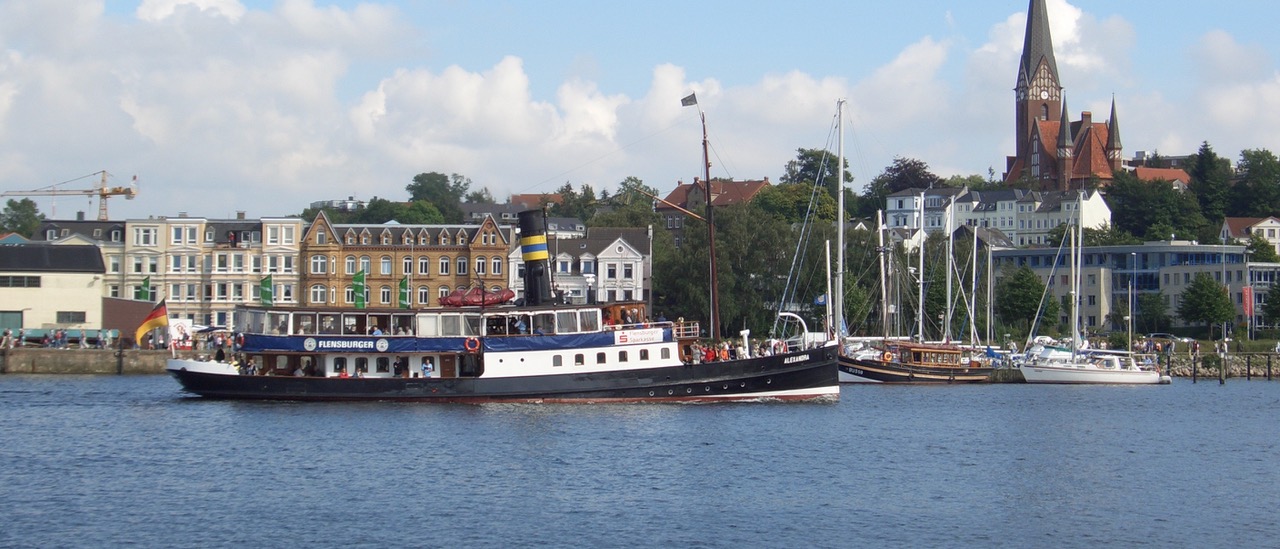 Blick von der Kaimauer am Westufer auf den Salondampfer Alexandra bei der Einfahrt in den Flensburger Hafen. Im Hintergrund ist das Ostufer mit dem Hafendamm und der markanten St. Jürgen Kirche zu sehen
