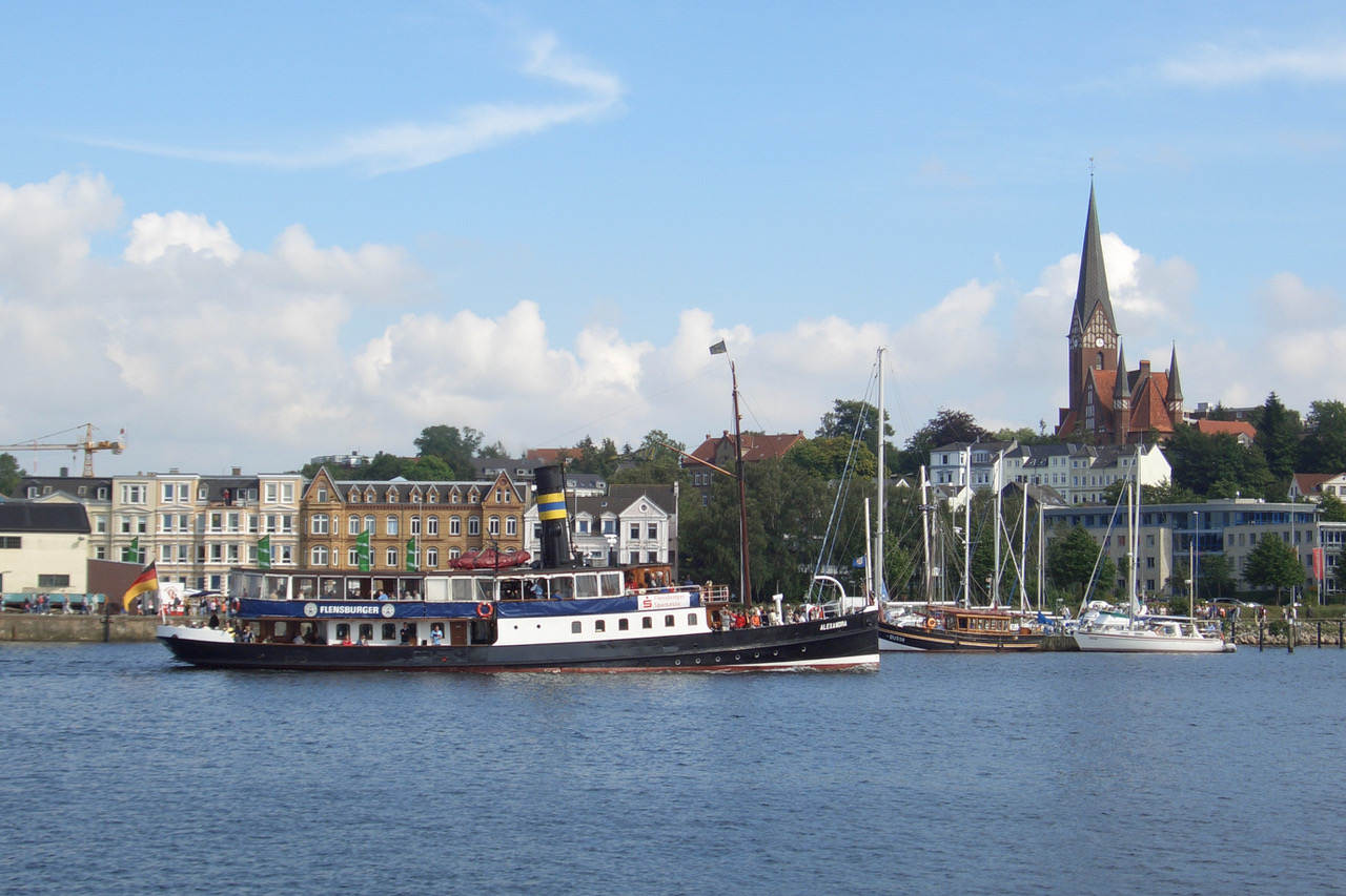 Blick von der Kaimauer am Westufer auf den Salondampfer Alexandra bei der Einfahrt in den Flensburger Hafen. Im Hintergrund ist das Ostufer mit dem Hafendamm und der markanten St. Jürgen Kirche zu sehen
