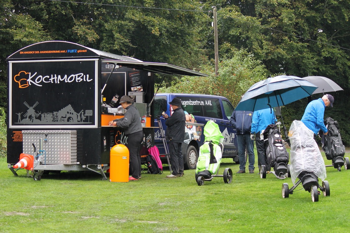 Fünf Golferinnen und Golfer stehen mit ihren Golfbags vor dem Kochmobil des AAK. Dort gibt es Bratwürste zur Stärkung