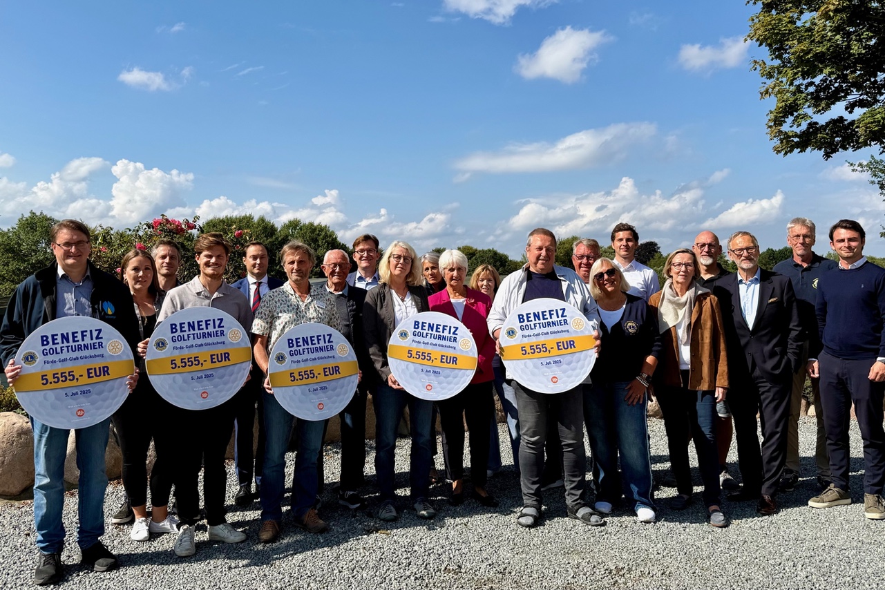 21 Personen stehen zum Gruppenfoto im Förde-Golfclub zusammen. 5 Personen halten dabei große, runde Schilder vor sich hin. Diese stellen Golfbälle da, die mit dem jeweiligen Spendenbetrag von 5.555 Euro beschriftet sind.