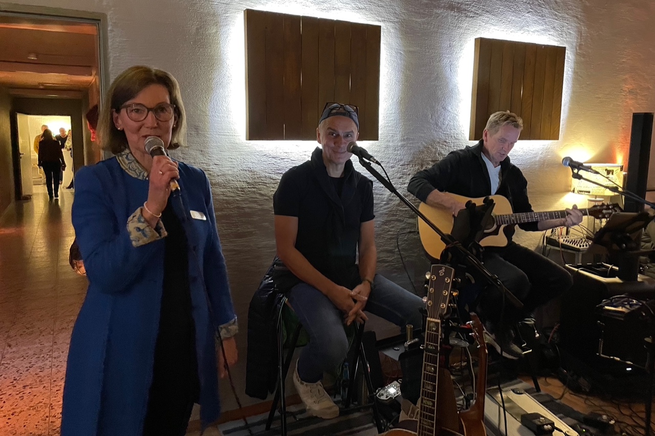 Inge-Marie Carstensen begrüßt die Besucher. Neben ihr die beiden Livemusiker mit Gitarren sitzend auf Barhockern.