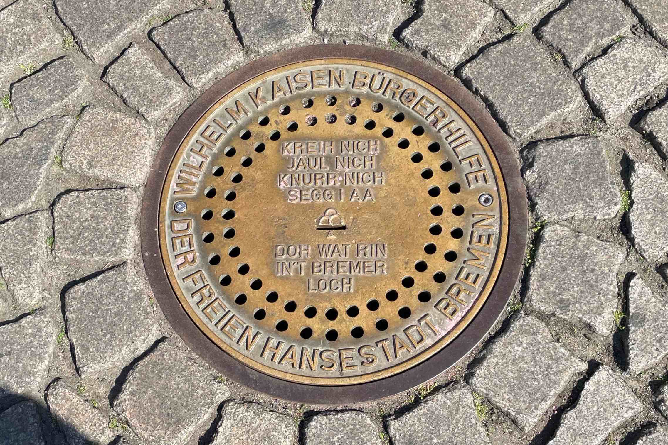 Beschrifteter Gullydeckel aus Bronze eingelassen im Basaltpflaster. Auf dem Deckel ist folgender Text: Im Außenkreis steht, Wilhelm Kaisen Bürgerhilfe der Freien Hansestadt Bremen. Über dem Einwurf-Schlitz in der Mitte steht kreih nich, jaul nich, knurr nich, segg I aa und darunter doh wat rin in´t Bremer Loch.